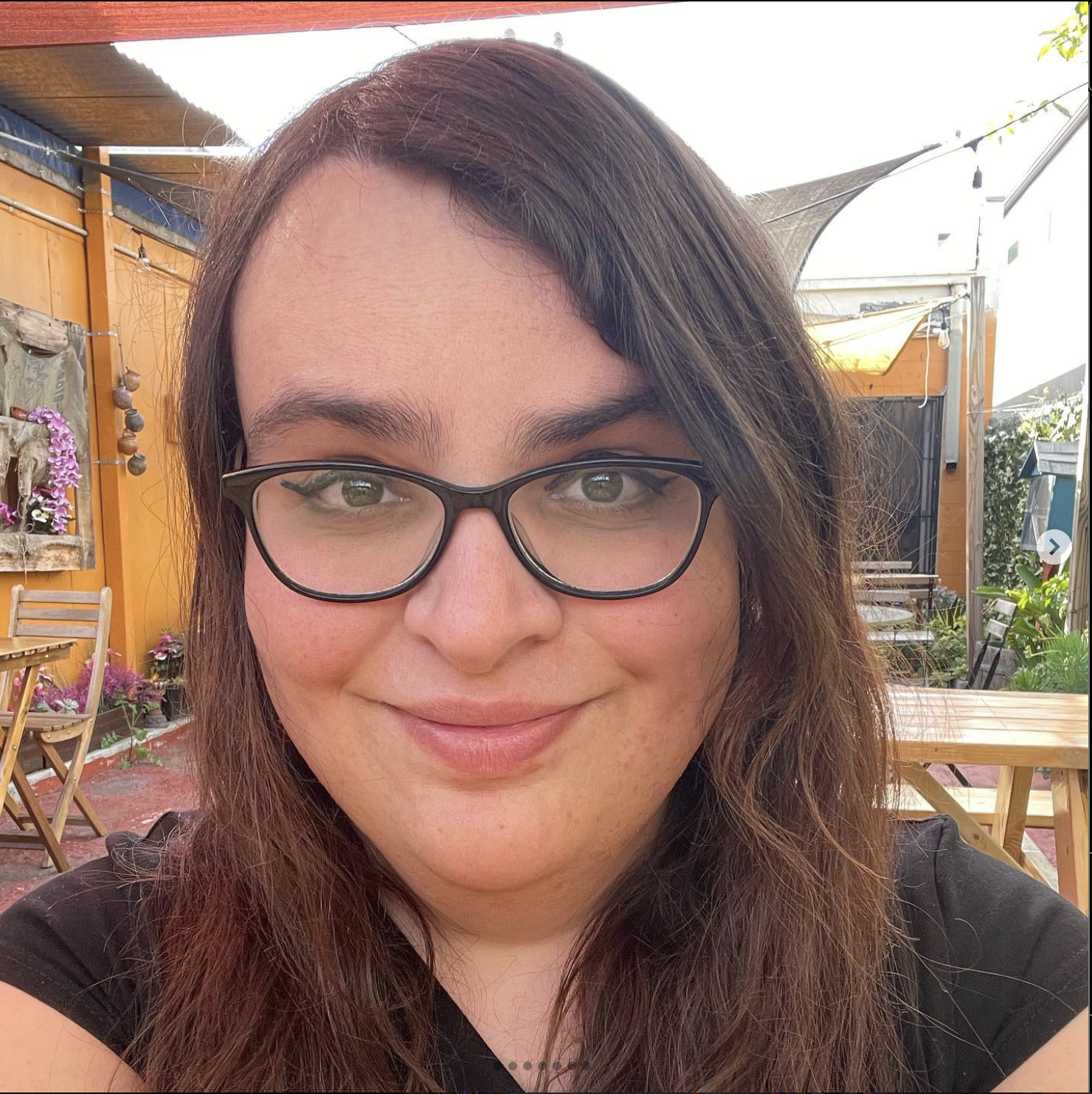 Chelsea Kelly-Reif, a software engineering leader with long brown hair and glasses smiles warmly outdoors, sitting in a cozy garden setting with wooden furniture, colorful flowers, and shaded awnings in the background.